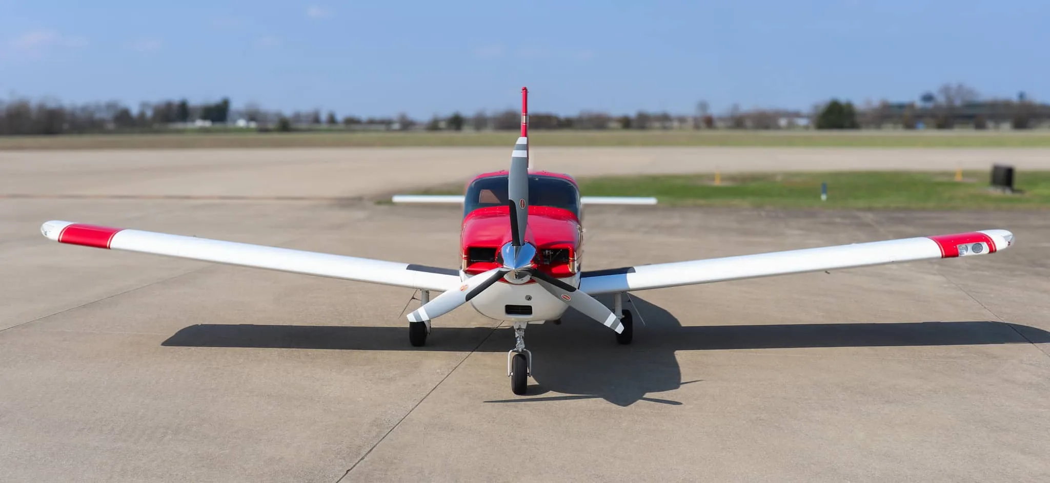 A red and white Socata Trinidad aircraft with gold stripes is parked on an airport tarmac, viewed directly from the front.