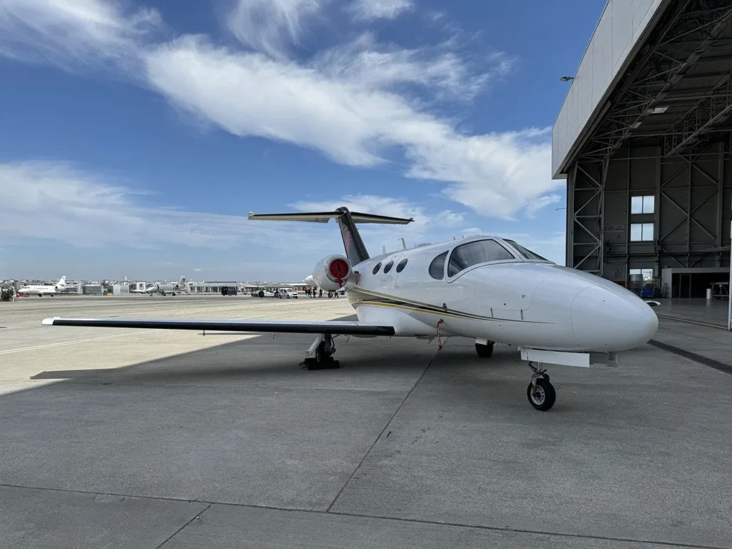 A white business jet with black and gold stripes on the tail, parked on an airport tarmac next to a large hangar.