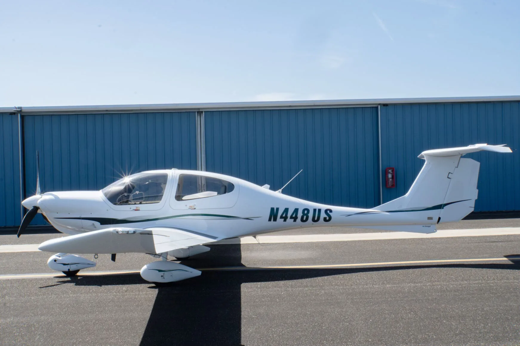 A white Diamond DA40 Star XL aircraft with green and grey stripes, registration N448US, parked on an tarmac in front of a blue hangar.
