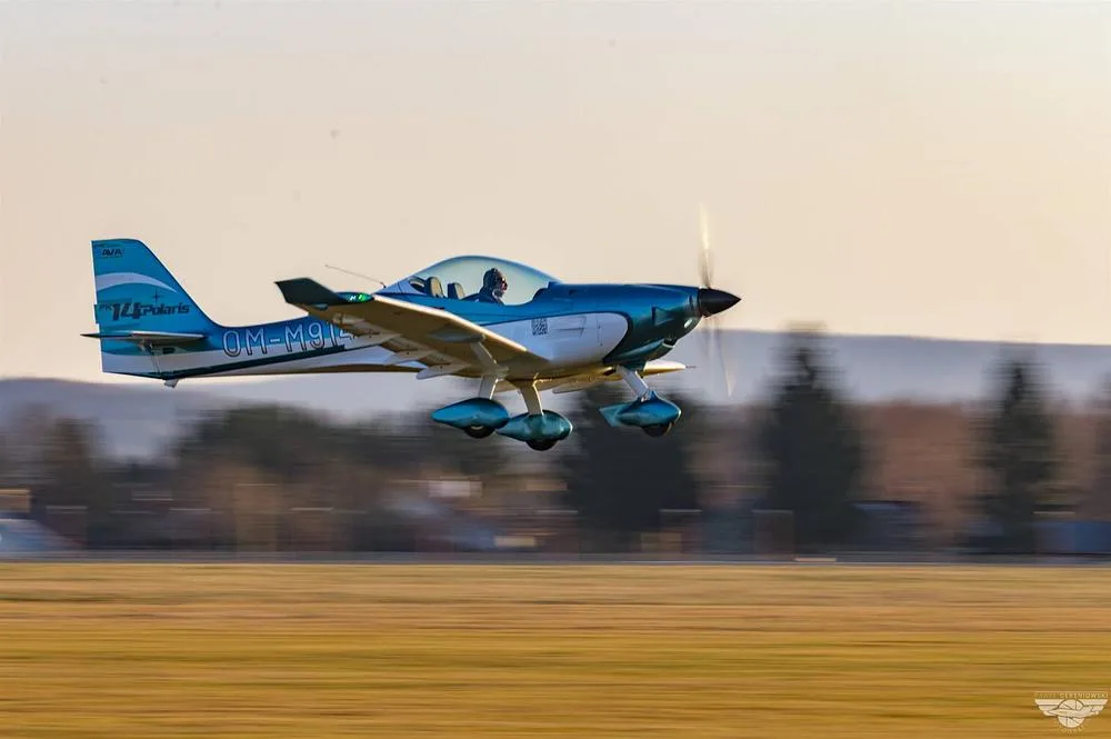 A blue and white FK14 Polaris aircraft (OM-M914) with a pilot, flying low over a blurred landscape.