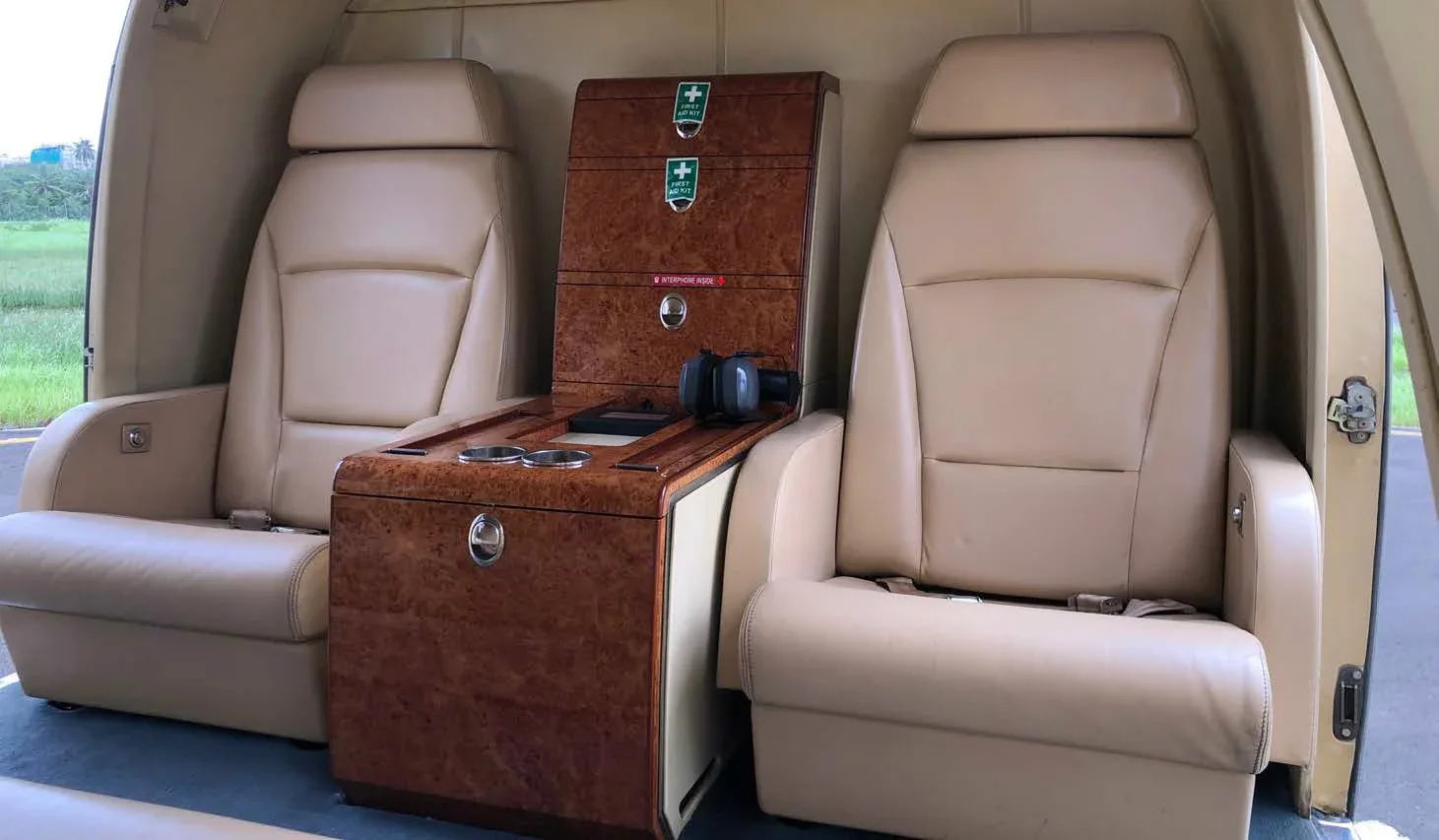 Two beige leather passenger seats facing forward with a wooden storage console between them inside a helicopter cabin.