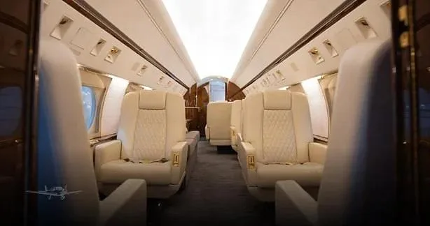 The spacious interior of a business jet cabin shows multiple cream-colored quilted leather seats along a central aisle.