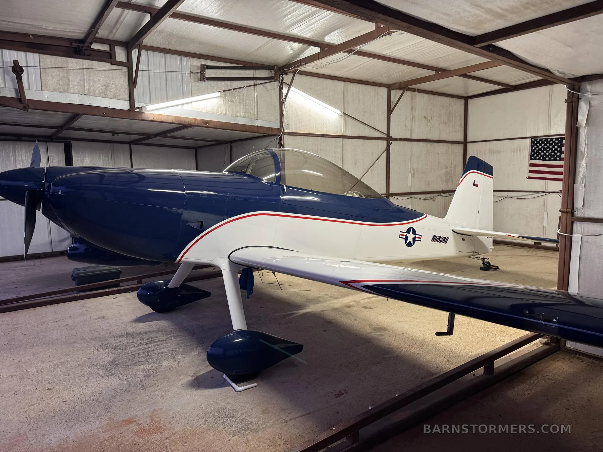 A blue and white single-engine aircraft, registration N663BH, parked inside a hangar with an American flag on the wall.