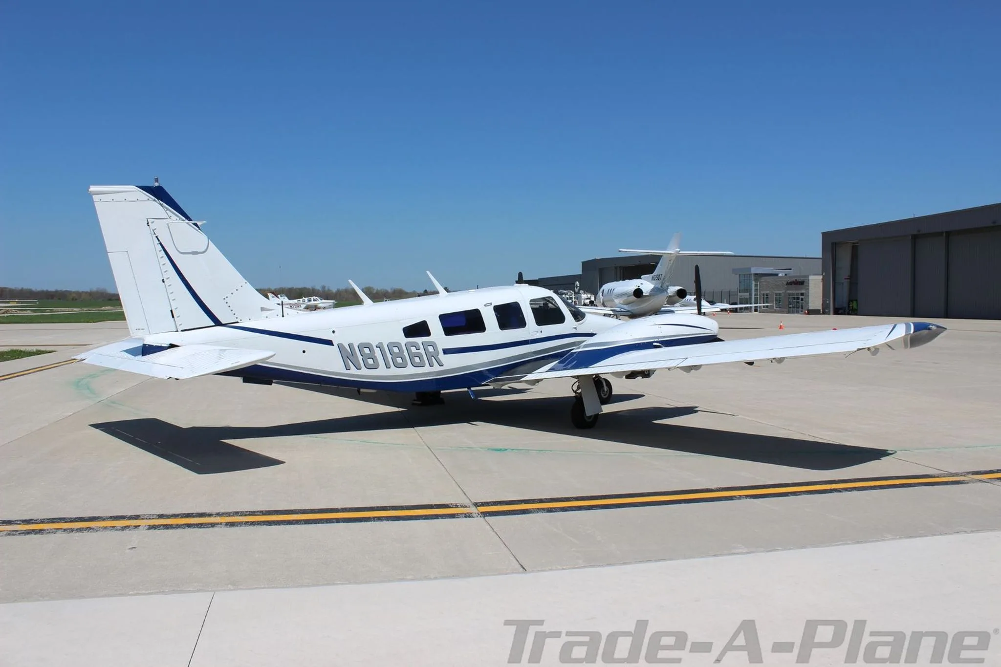 A rear-quarter view of the white and blue Piper Seneca aircraft N8186R parked on an airport tarmac with hangars in the distance.