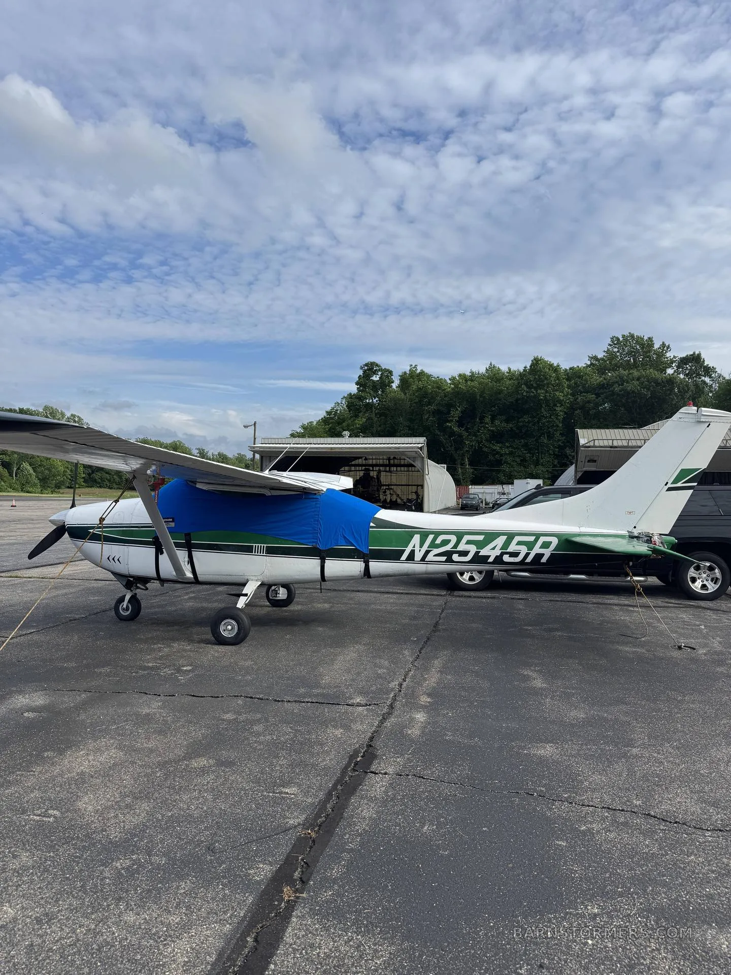 A white and green single-engine propeller aircraft with a blue engine cover is parked on an asphalt ramp, viewed from the right side.