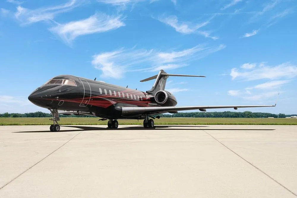 A black and red Bombardier Global 7500 business jet is parked on a tarmac under a blue sky with wispy clouds.