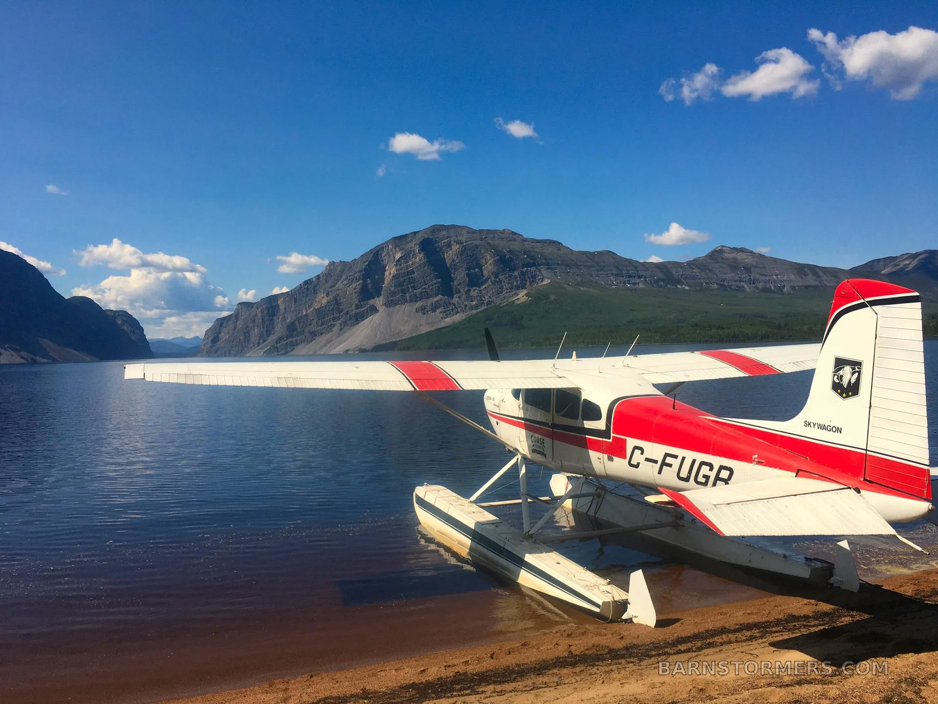 A white and red Cessna 180 (C-FUGR) on floats is partially in a lake with mountains and a blue sky in the background.