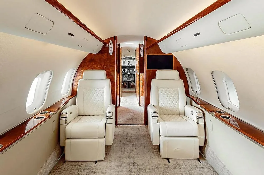 A view down the aisle of a private jet cabin, showing two white leather seats in the foreground and the cockpit entrance beyond.