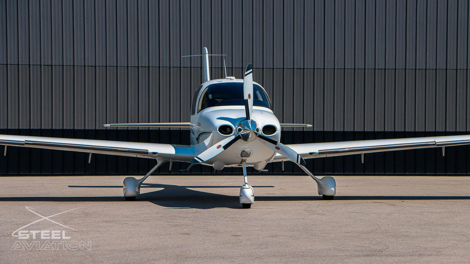 Close-up front view of a white Cirrus SR22 aircraft, highlighting its propeller and landing gear, in front of a grey hangar.
