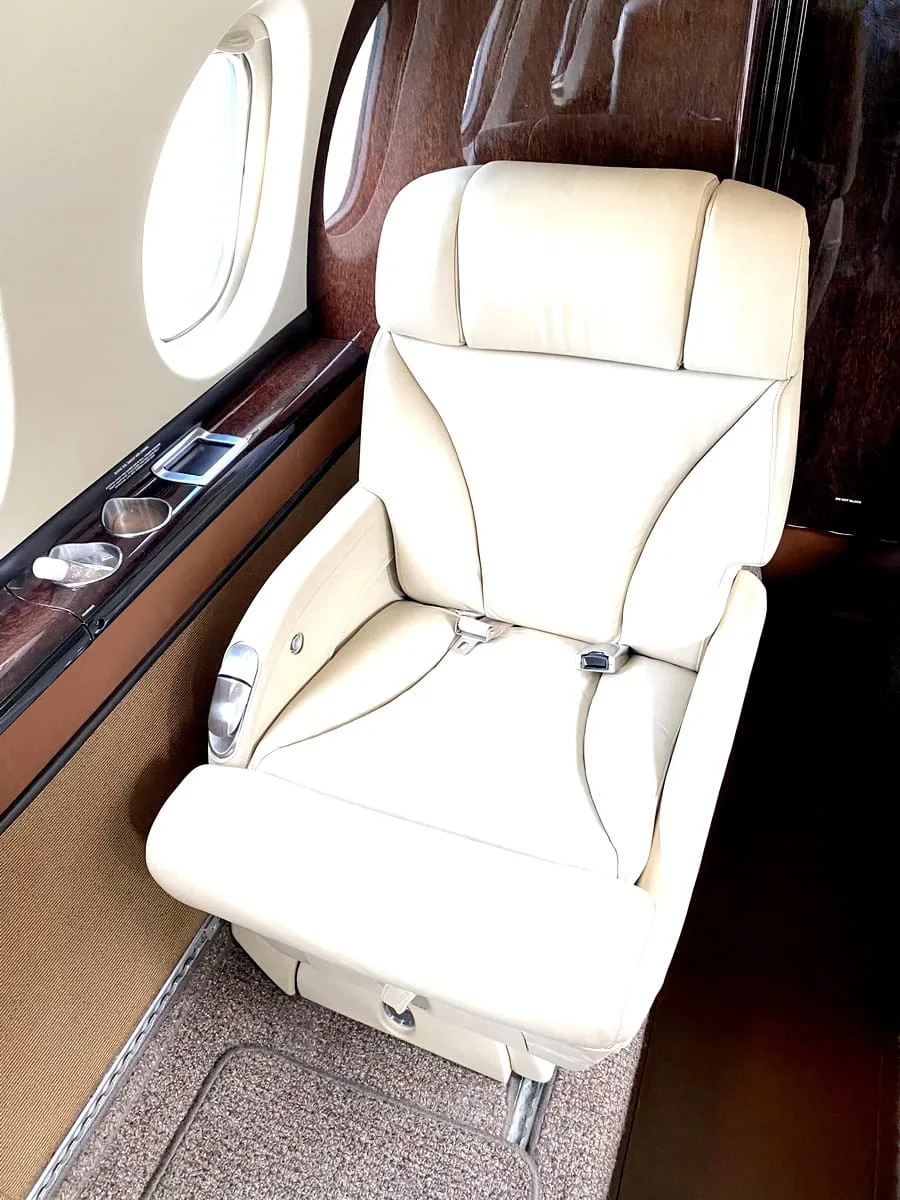 A luxurious cream-colored leather seat with a seatbelt in an aircraft cabin, next to a window.
