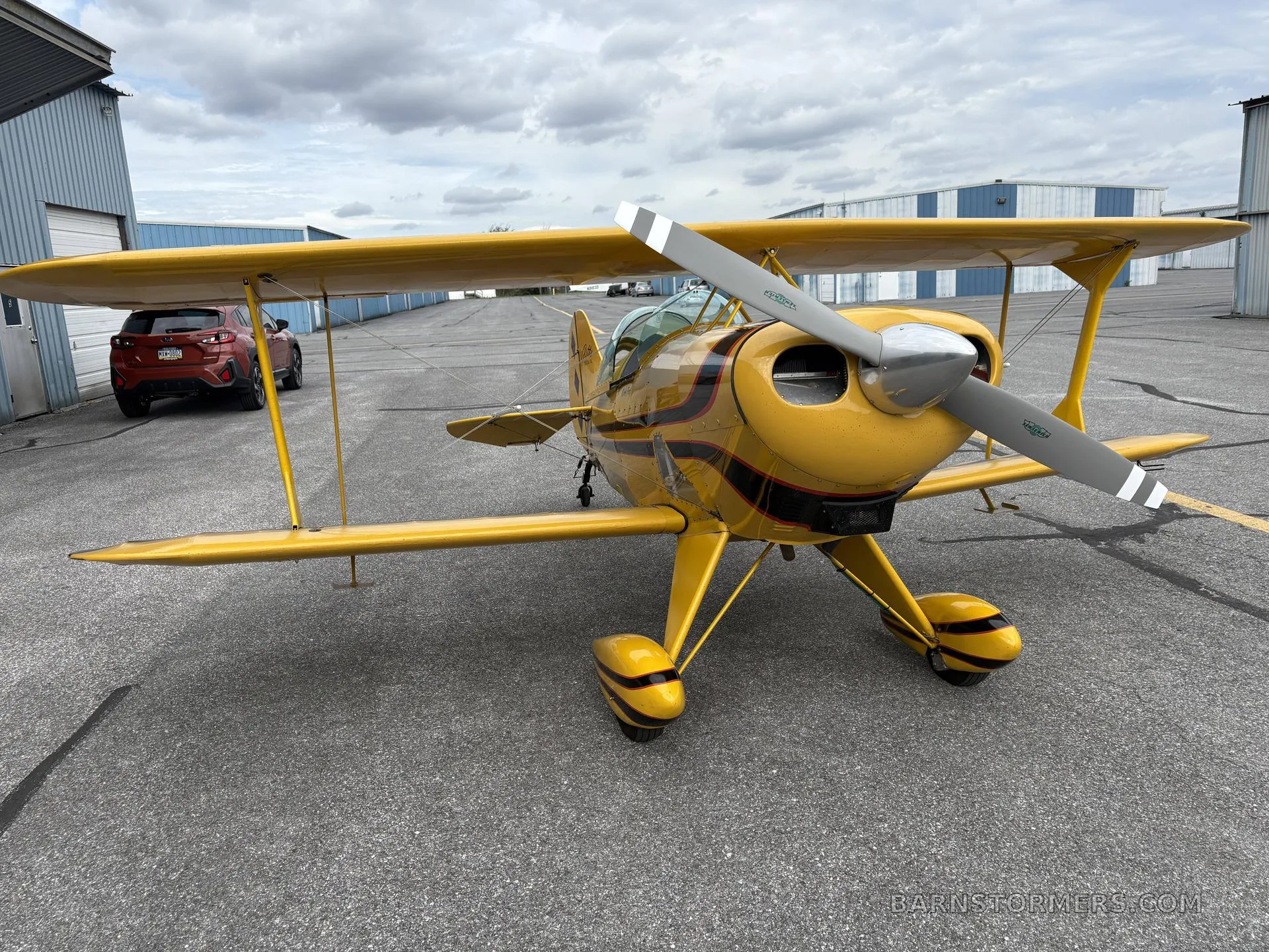 A yellow biplane with black and red stripes on the fuselage, parked on an asphalt apron with hangars in the background.