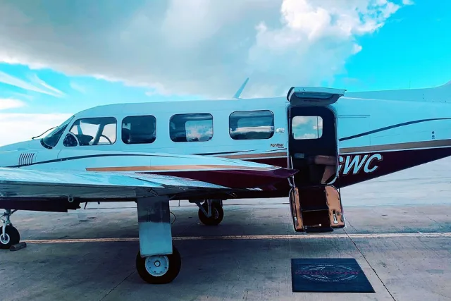 A white and maroon Piper PA-46 Malibu/Mirage aircraft with an open passenger door, parked on an tarmac under a partly cloudy sky.