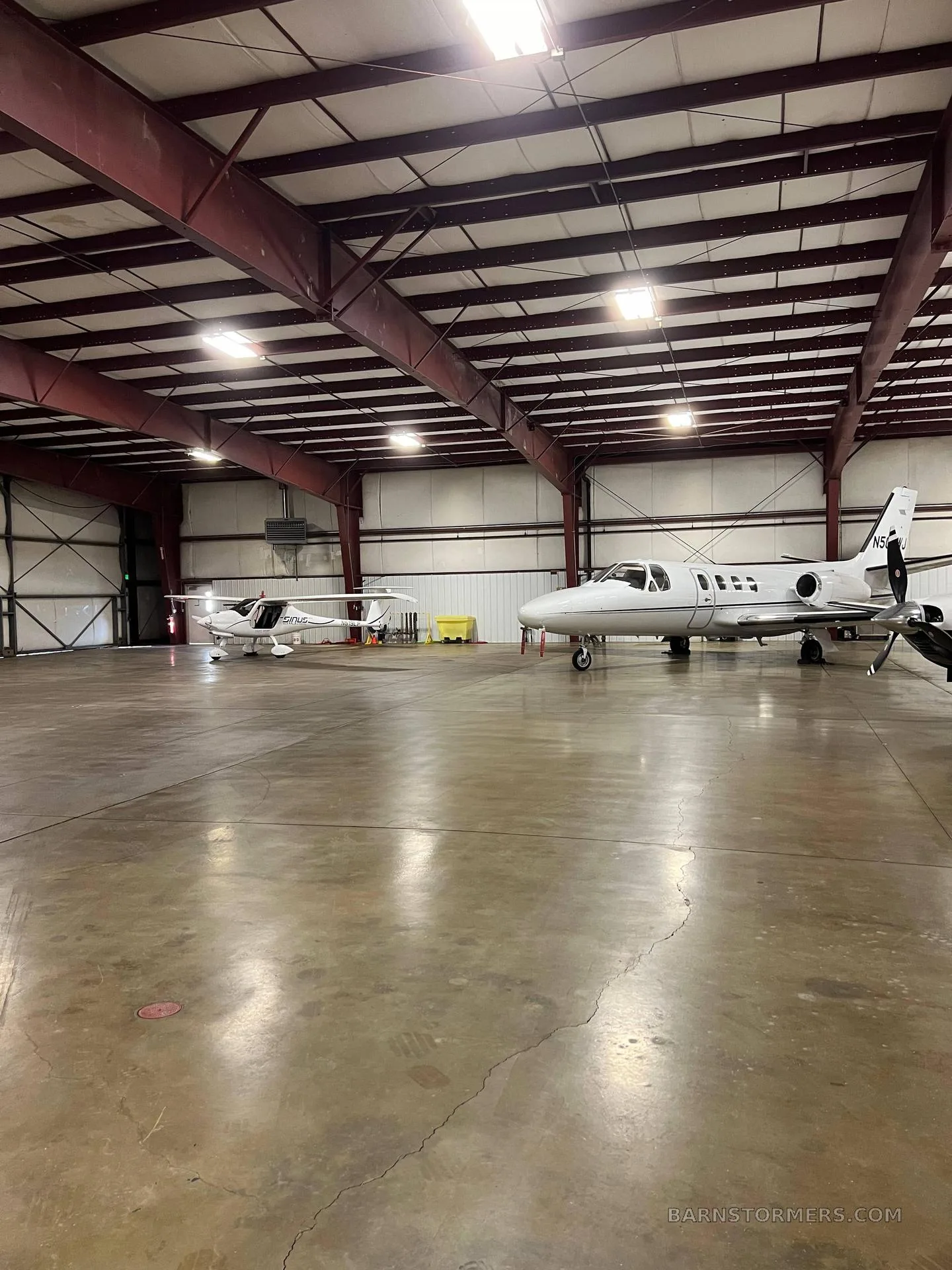 A white light sport aircraft and a white business jet are parked inside a spacious hangar with a concrete floor.