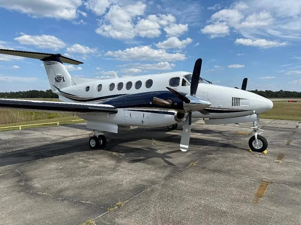 A side view of the white and dark blue Beechcraft King Air N3PX on an tarmac with a runway in the background.