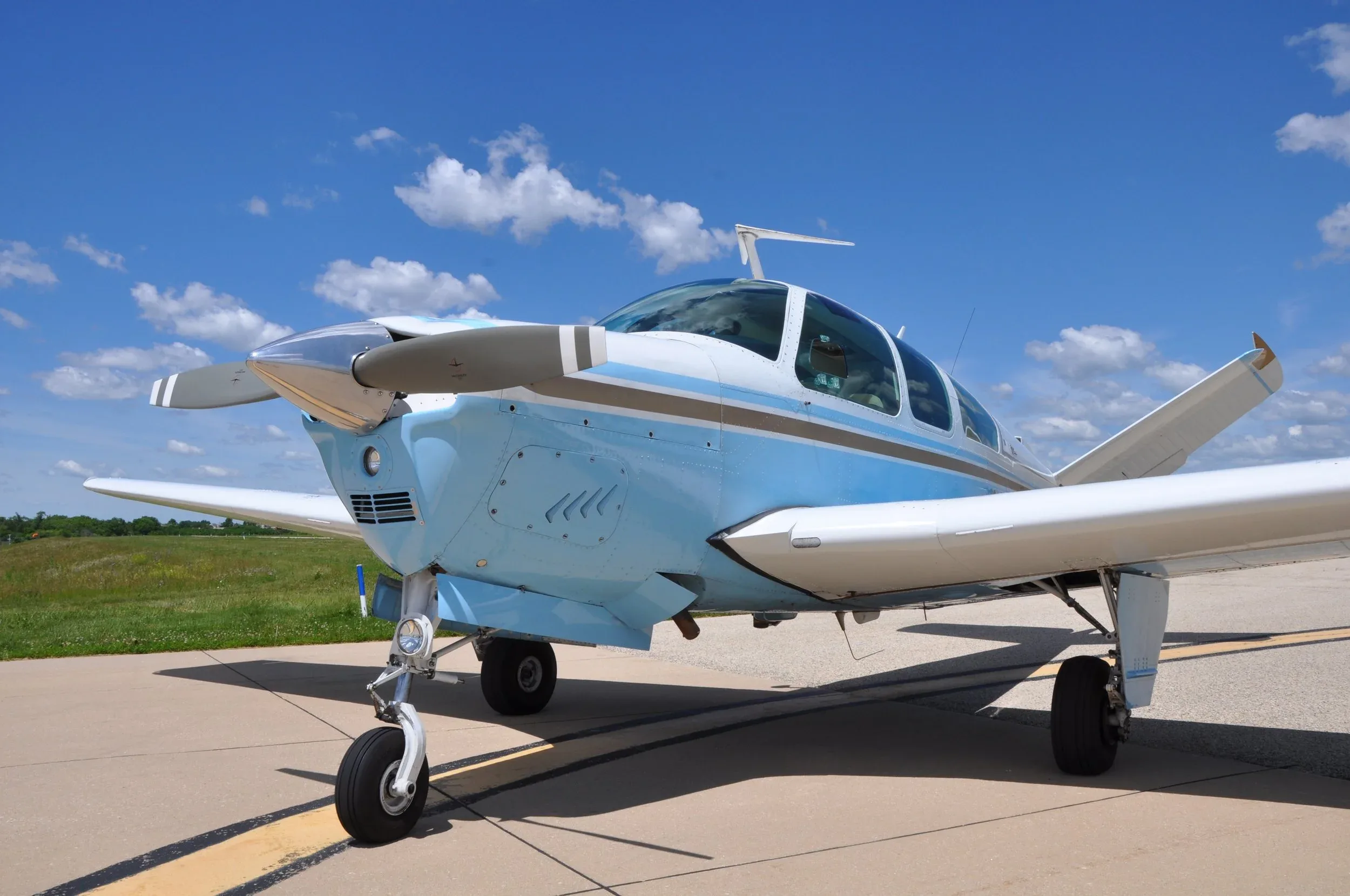 A close-up, low-angle view of the front of a light blue and white single-engine aircraft with a V-tail on an asphalt runway.