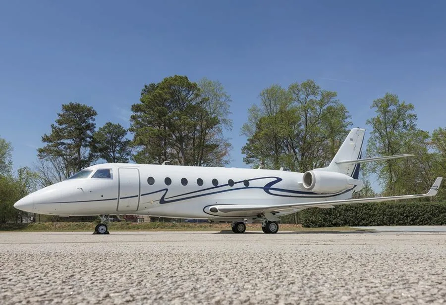 A white business jet with a distinctive blue wavy stripe on its fuselage, parked on an airfield under a clear blue sky.