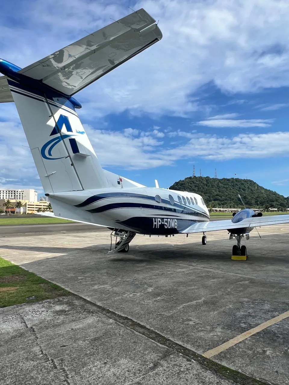 A white and blue turboprop aircraft (King Air) with registration HP-5DMB, viewed from the rear, parked on an airport tarmac with a green hill in the background.