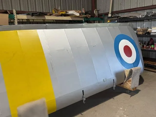 A close-up of the silver fuselage side of a biplane, featuring a yellow stripe and a red, white, and blue roundel.