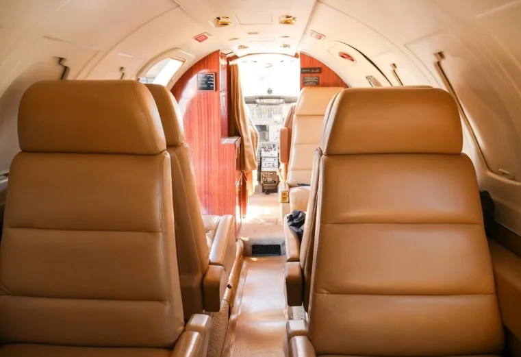 A view down the aisle of a private jet cabin, showing multiple rows of tan leather seats and wood paneling.