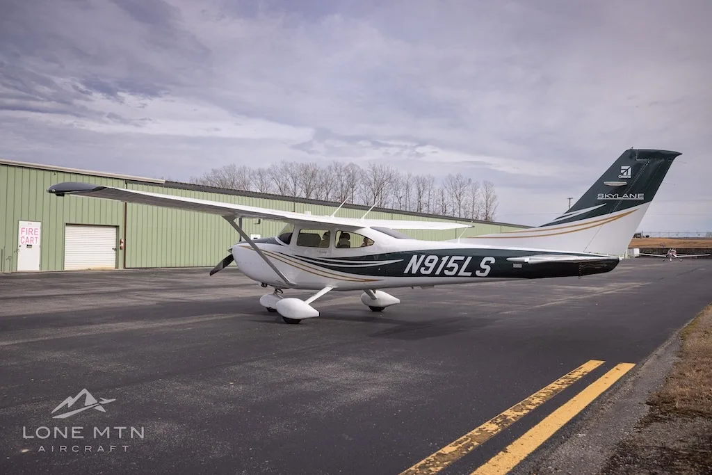 A side view of a white and dark green Cessna Skylane N915LS with gold stripes, parked on an asphalt ramp.