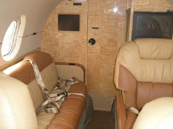 A close-up view of the private jet interior, showing a tan and brown leather seat with a seatbelt and a wooden bulkhead.