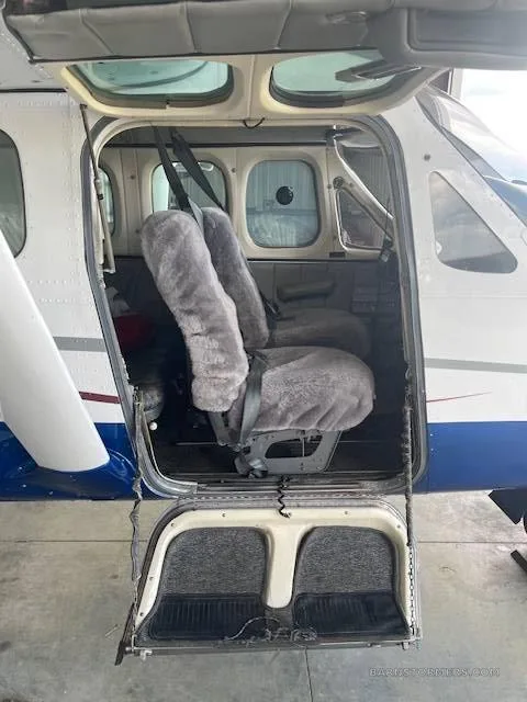 The open cabin door of a white and blue aircraft, revealing two light gray fuzzy-covered seats and the step-down entry.