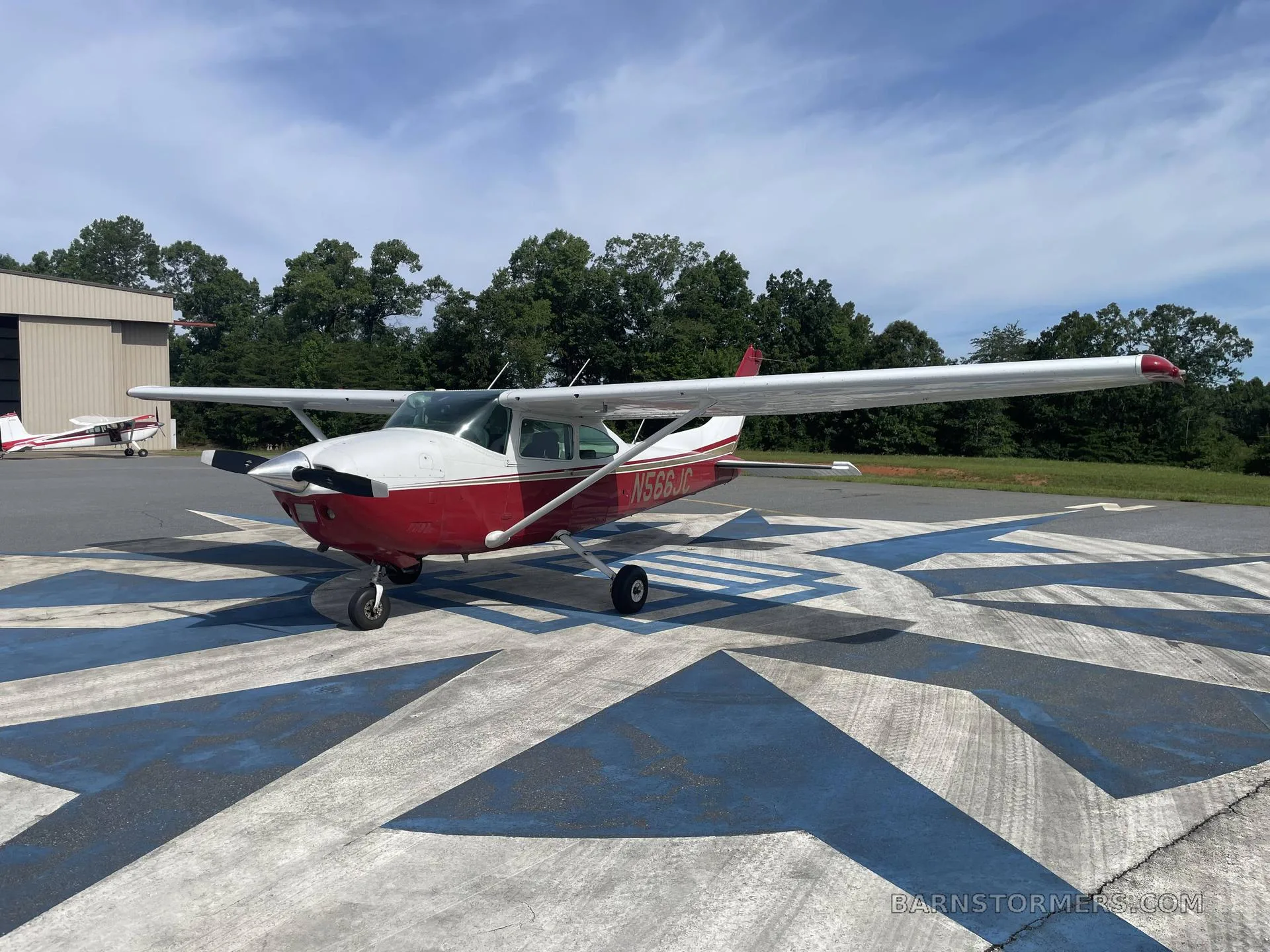 A red and white Cessna N566JC aircraft is parked on a patterned concrete pad with a hangar and trees in the background.