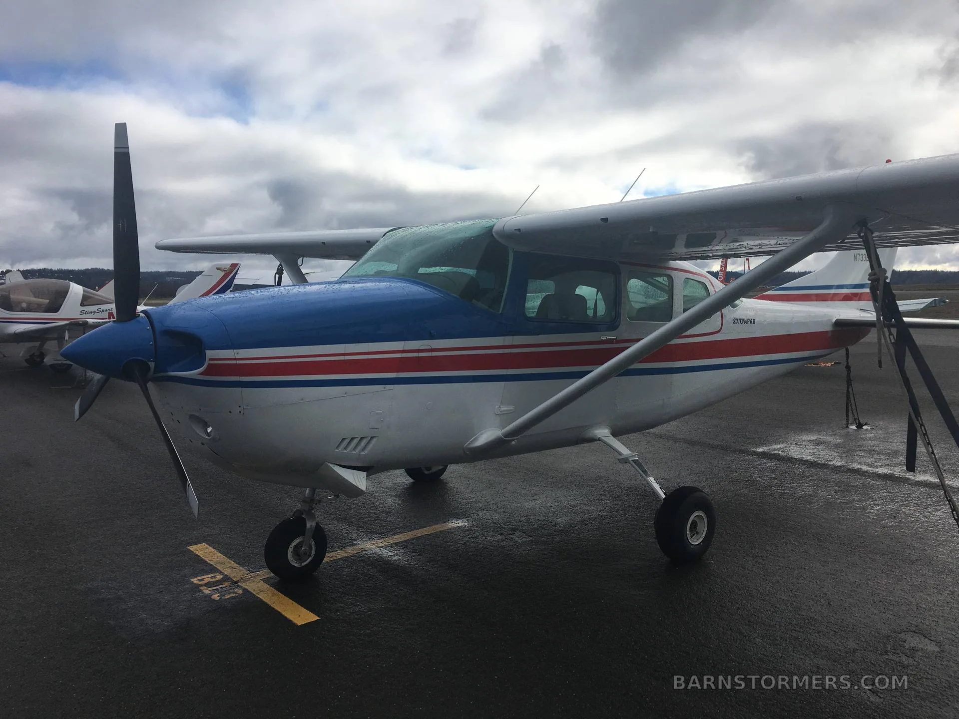 A three-quarter front view of a white, red, and blue Cessna Stationair 6 II (N733EH) parked on a wet tarmac.