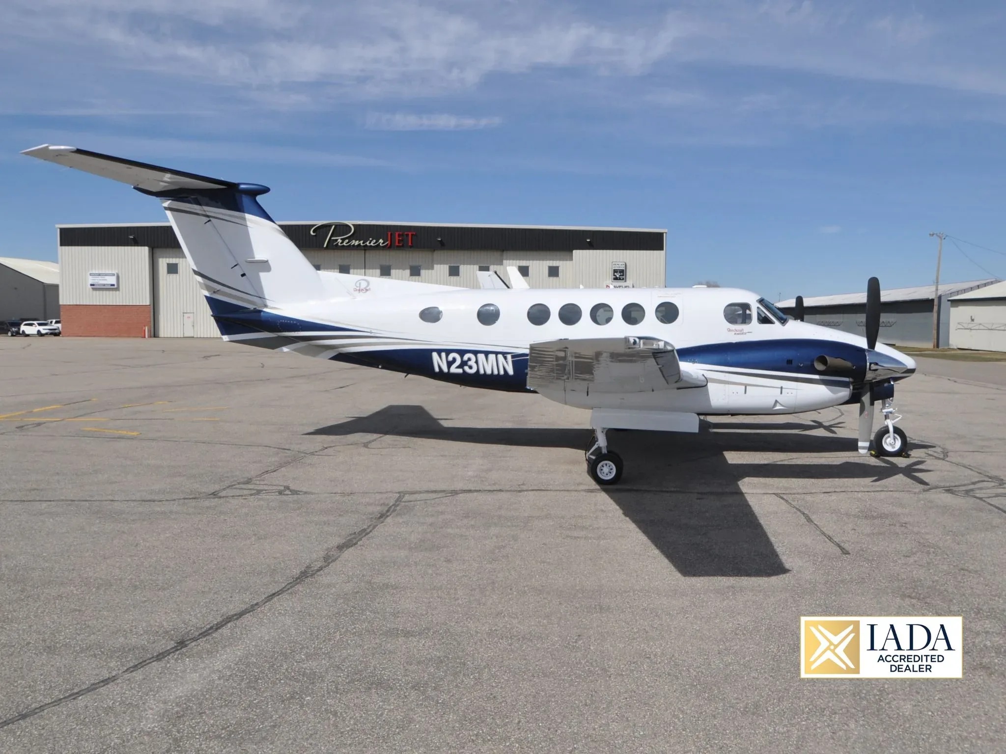 A white and blue twin-turboprop aircraft, likely a Beechcraft King Air, parked on an airport apron with hangars in the background.