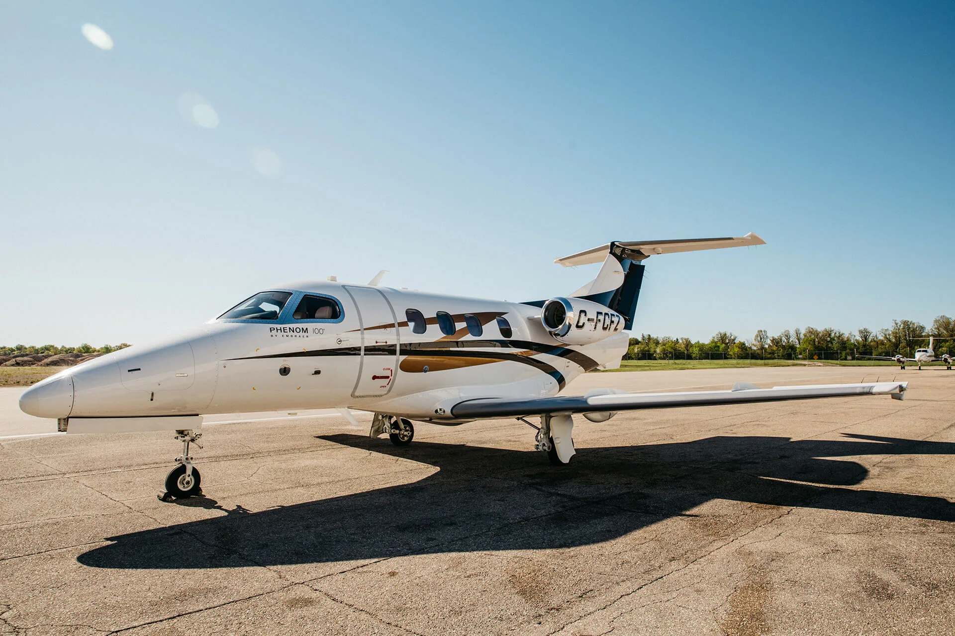 A white Embraer Phenom 100 business jet with blue and gold stripes is parked on an asphalt tarmac on a sunny day.