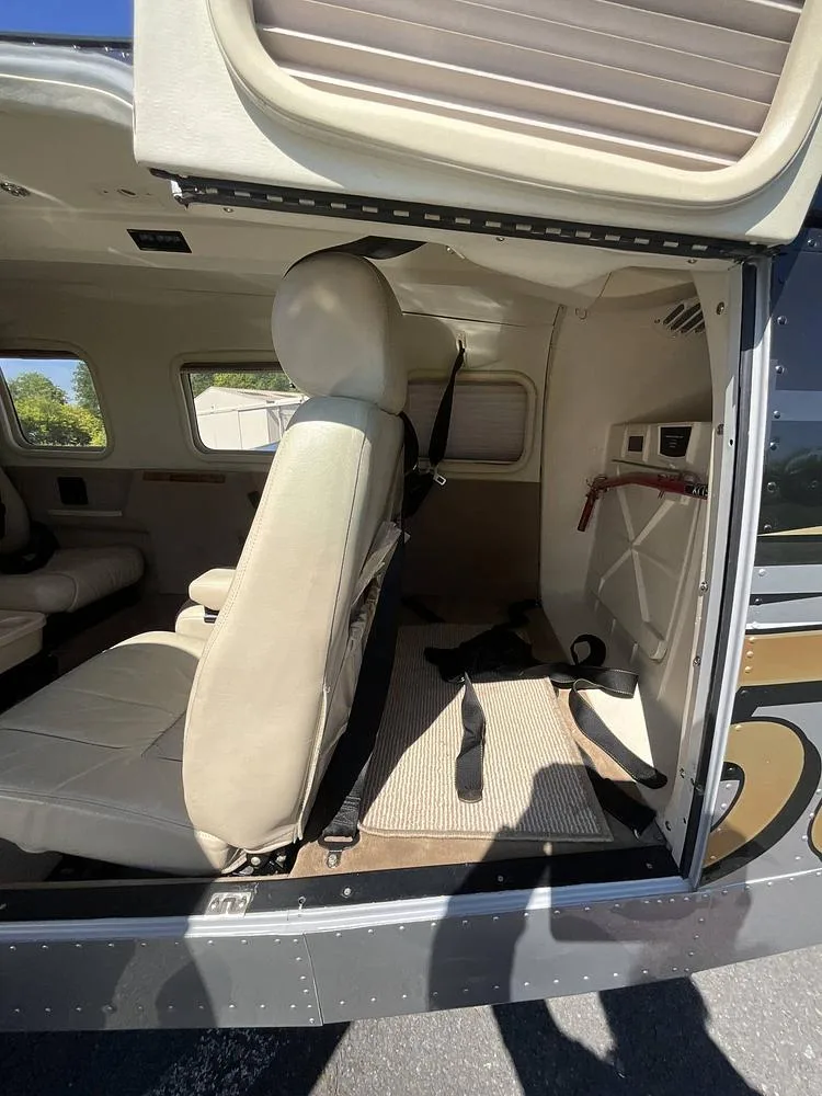 View into the open door of a Piper Saratoga, showing a beige leather passenger seat and the rear cargo area.