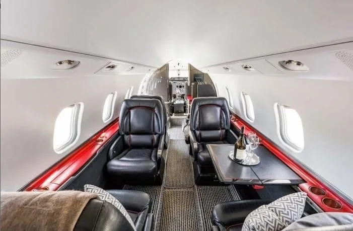 The spacious interior cabin of a private jet featuring black leather seats and a table with a wine bottle.