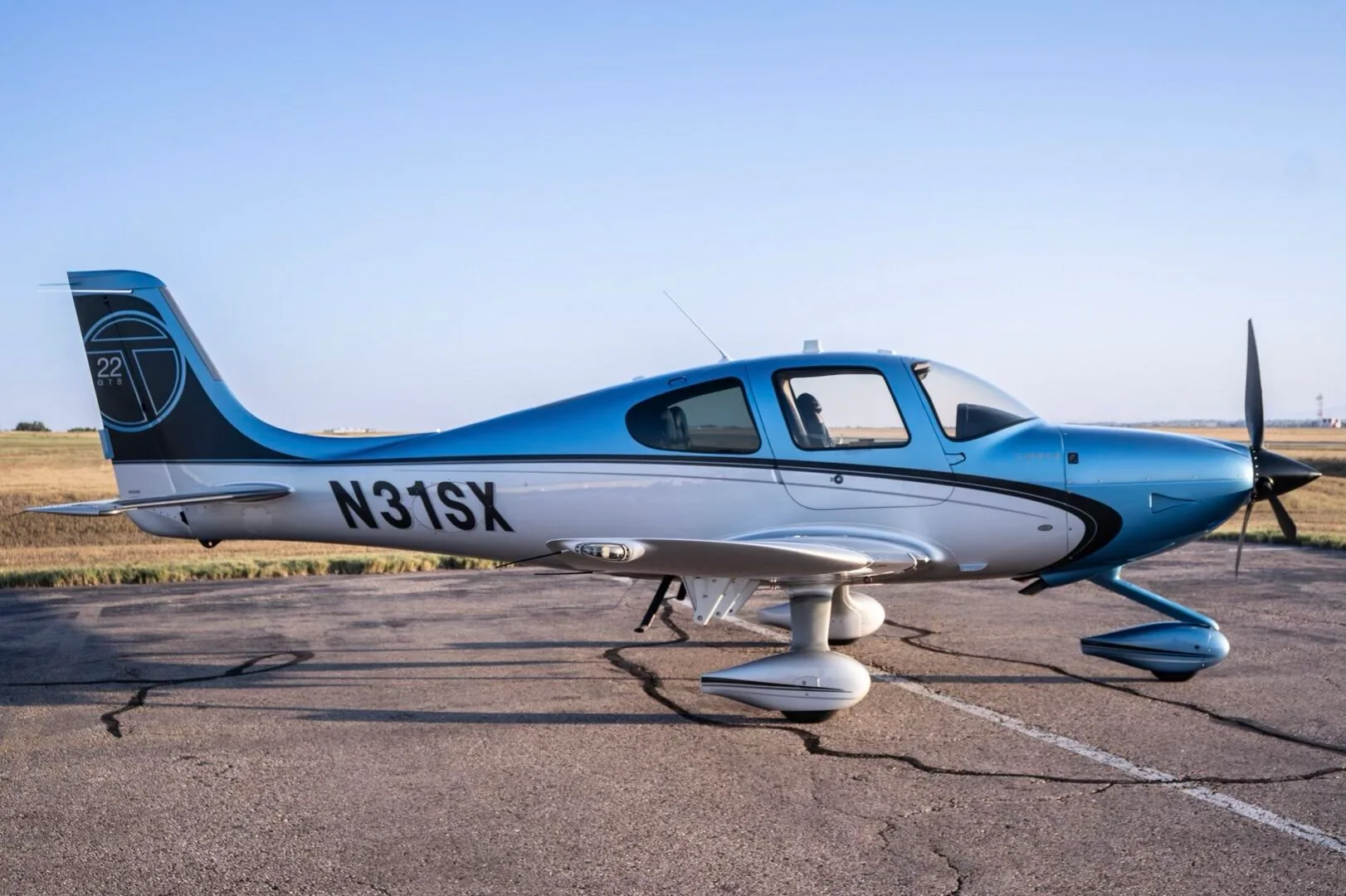A Cirrus SR22T aircraft, N31SX, in blue and silver paint, parked on an airfield with a clear sky.