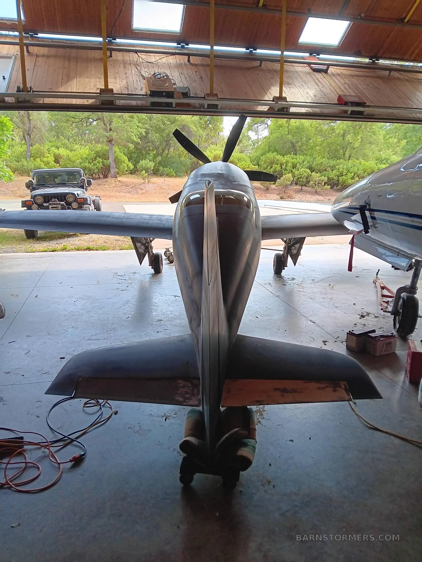 A dark, single-engine aircraft with a tailwheel configuration, viewed from the rear, inside a hangar with the door open to a wooded area.
