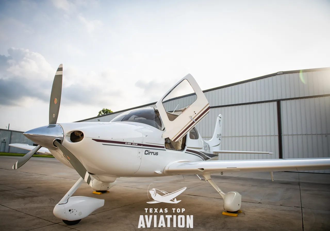 A white Cirrus SR22 aircraft with maroon and gold stripes is parked on a tarmac with its passenger door open, under a cloudy sky.
