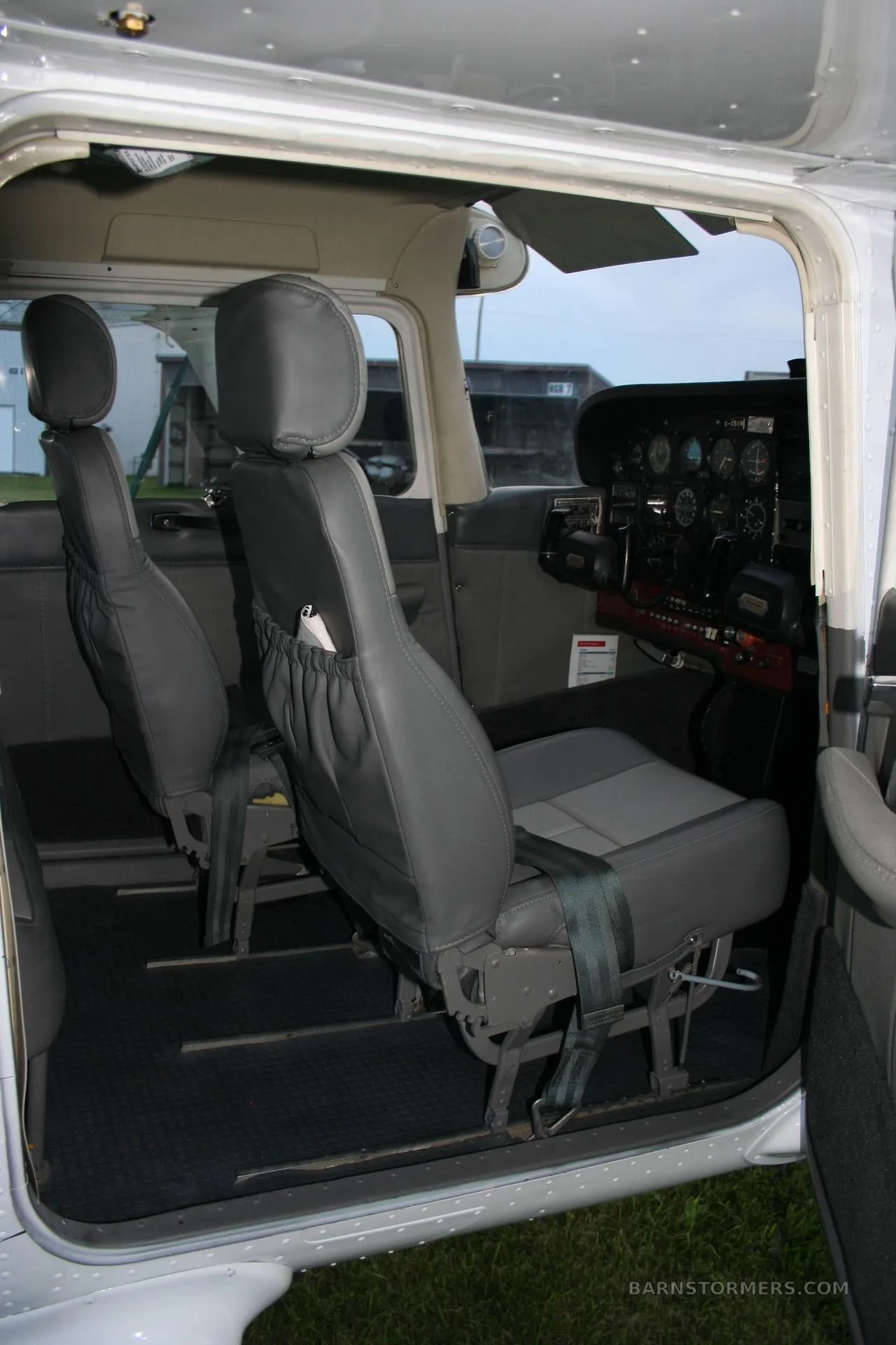 The gray and light gray front seats of a small aircraft cabin are visible, with a partial view of the cockpit and outside.