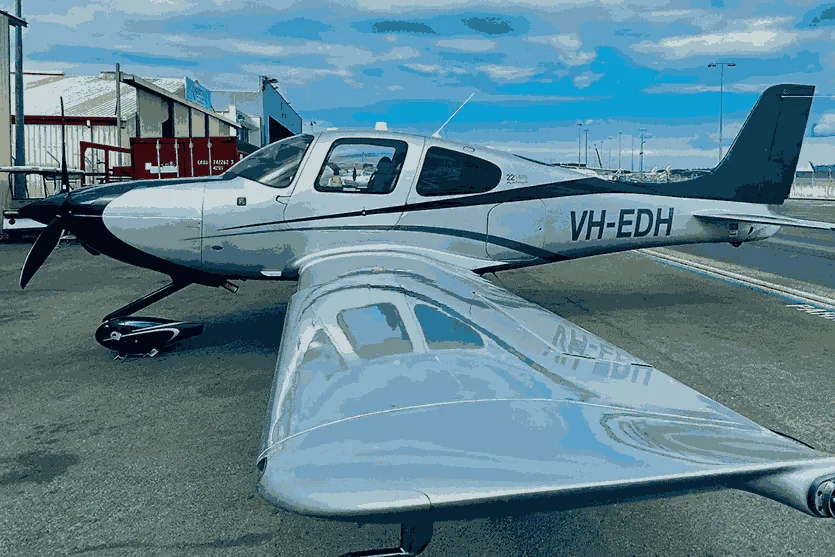 A silver and black single-engine propeller aircraft, registration VH-EDH, parked on an airfield with hangars in the background.