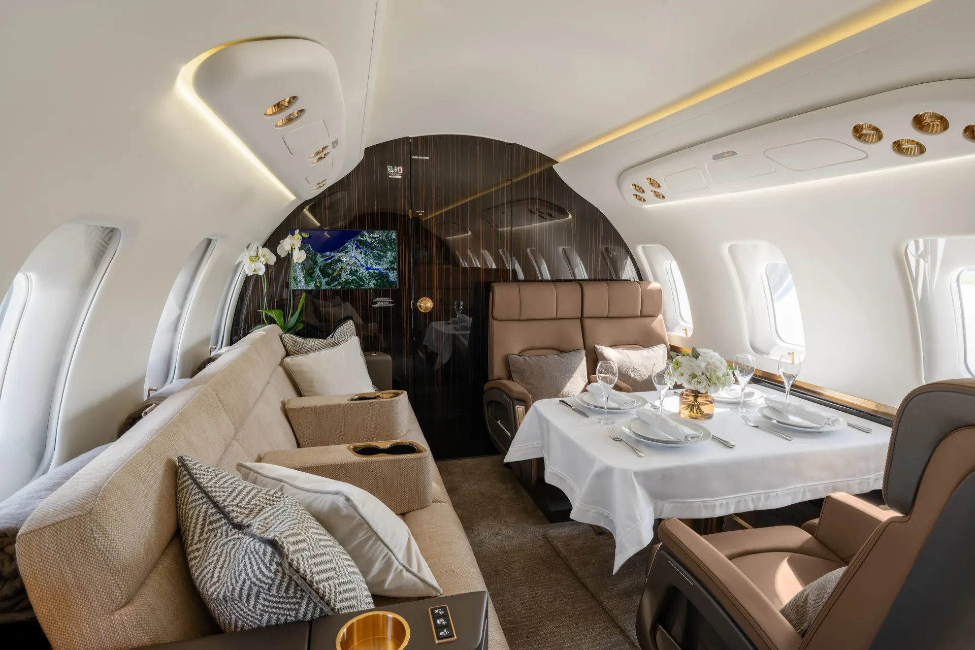 A luxurious aircraft cabin interior featuring a dining table set for four, a beige sofa, and brown leather seats.
