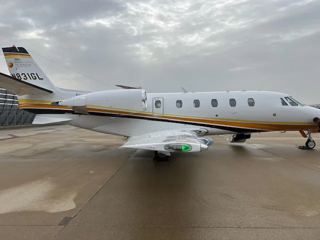 The N831GL business jet with orange and black stripes, parked on a wet tarmac under a cloudy sky.