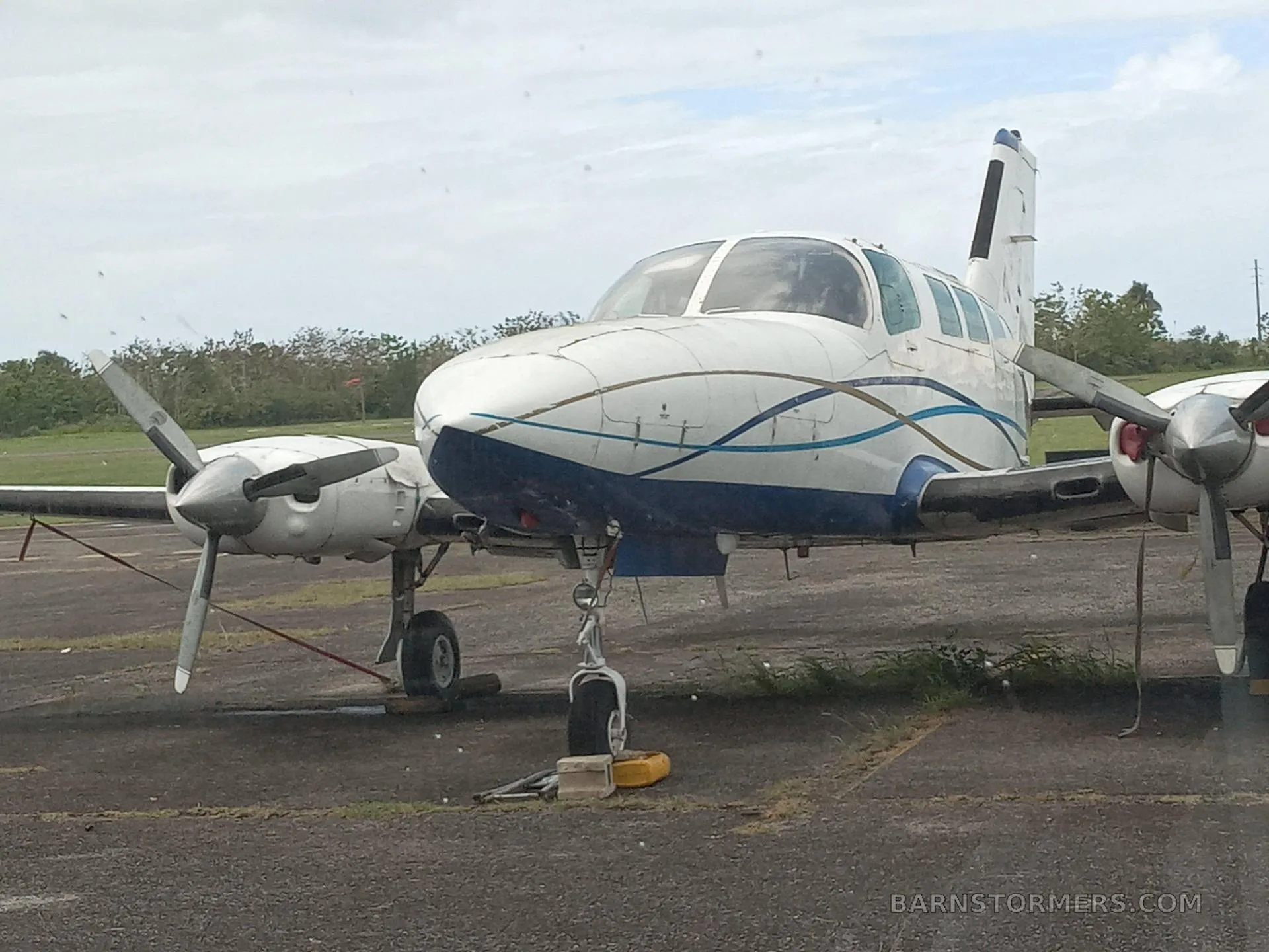 A white and blue twin-engine propeller aircraft with a blue stripe design, parked on an asphalt surface under an overcast sky.