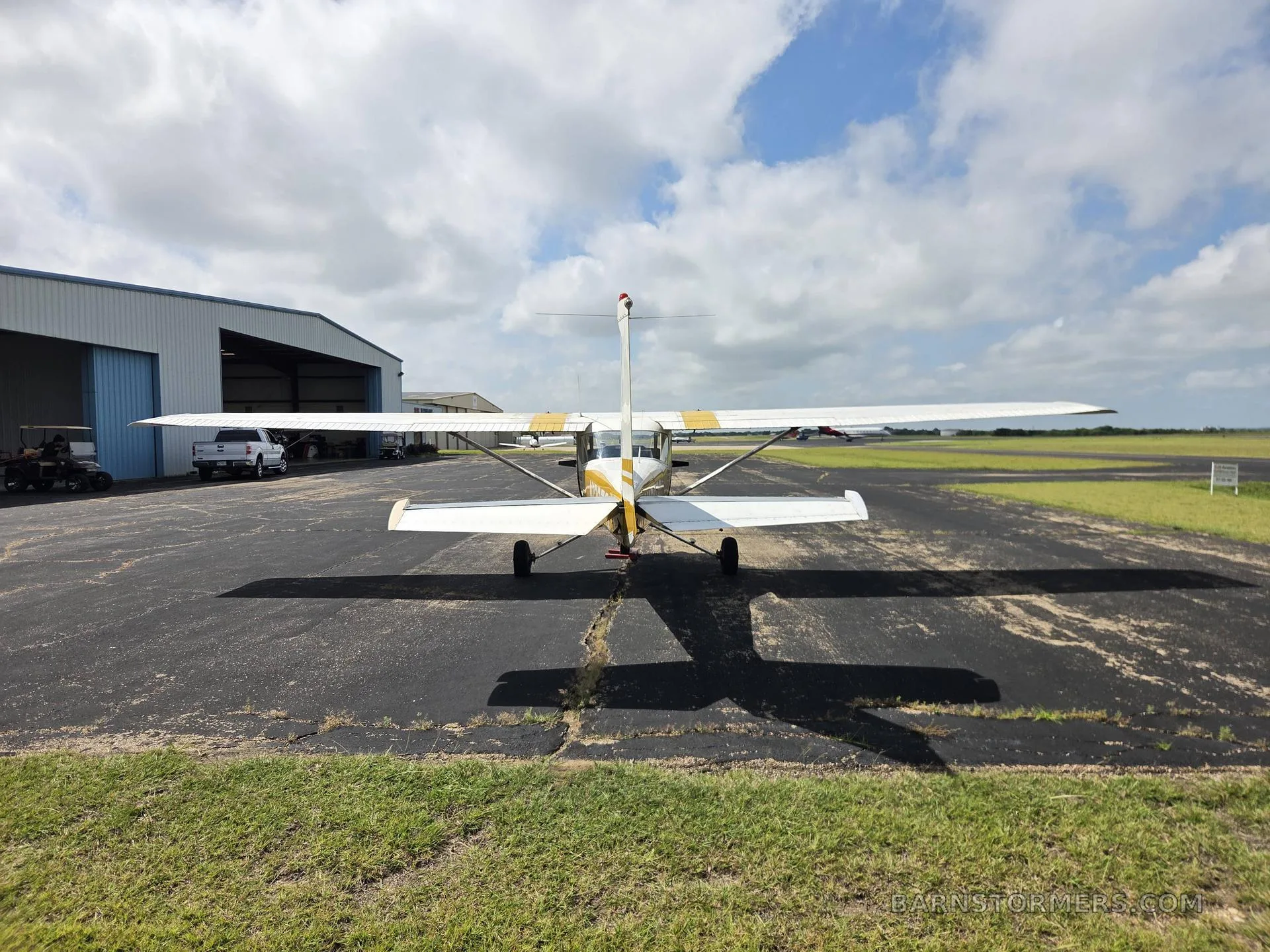A white and yellow light aircraft (N13370) parked on an asphalt ramp, viewed from directly behind, with a hangar in the background.