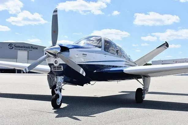 A front view of a white and blue single-engine propeller aircraft on an airfield under a partly cloudy sky.