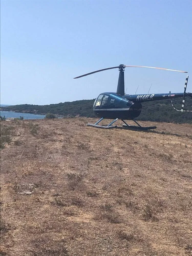 A dark blue Robinson R44 Clipper II helicopter, N144LR, is parked on a dry, grassy hill overlooking the sea.