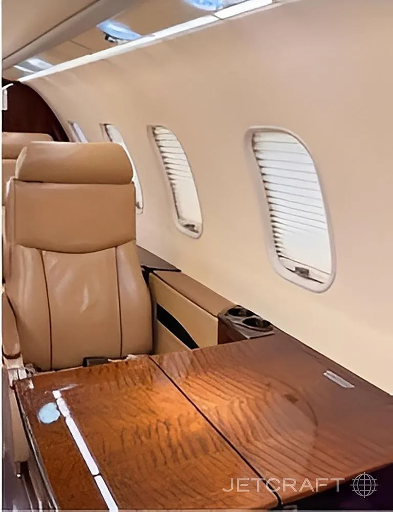 A close-up view of a tan leather seat and a polished wooden table next to windows with blinds in an aircraft cabin.