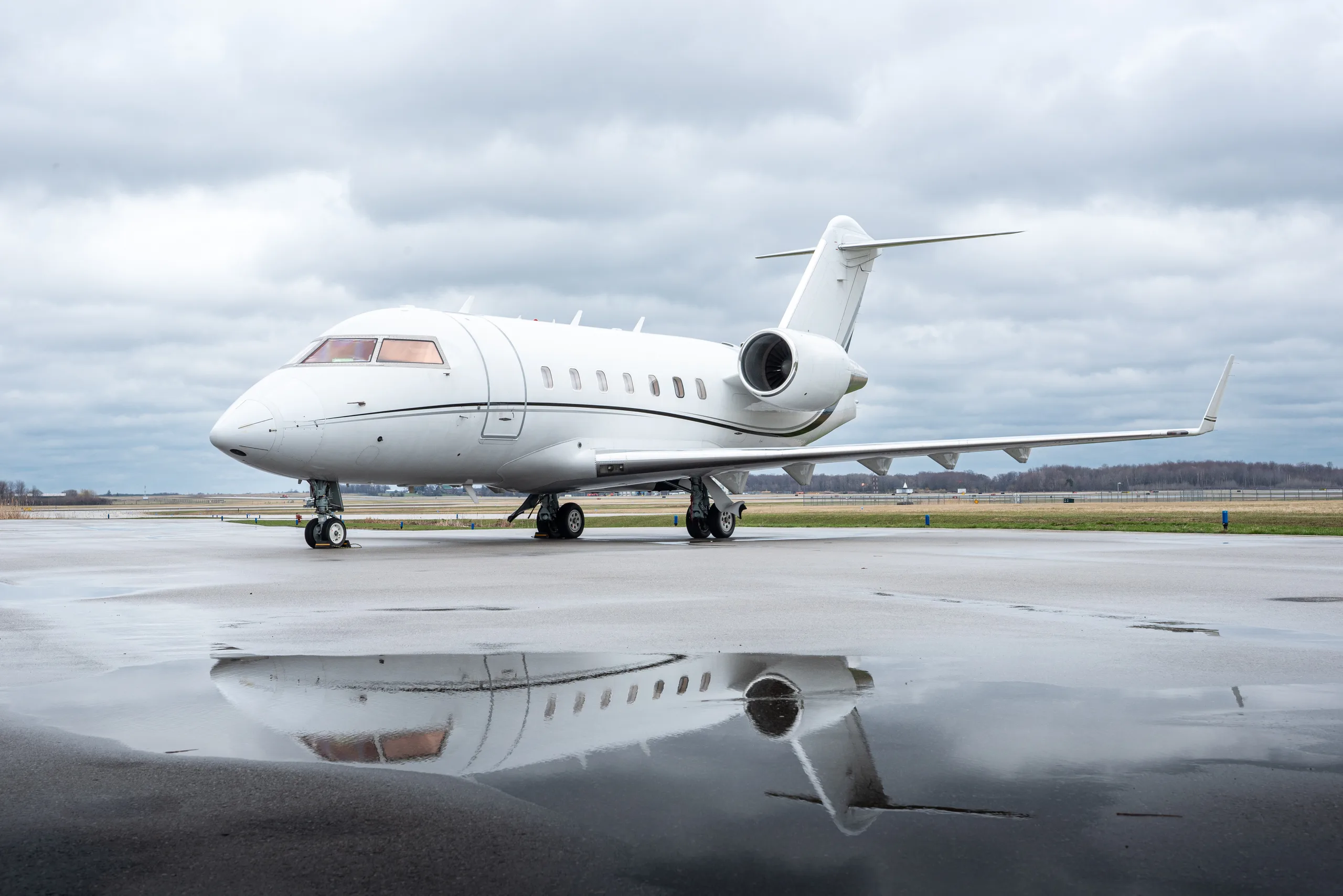 A white business jet with a dark stripe on a wet tarmac under a cloudy sky.