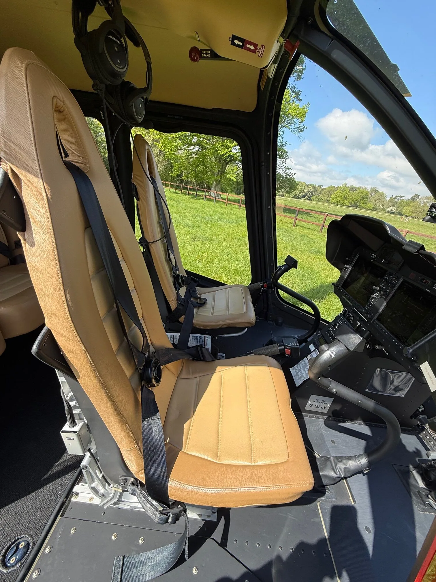 The tan leather pilot's seat and instrument panel of a Bell 505 helicopter with a green field visible through the windshield.