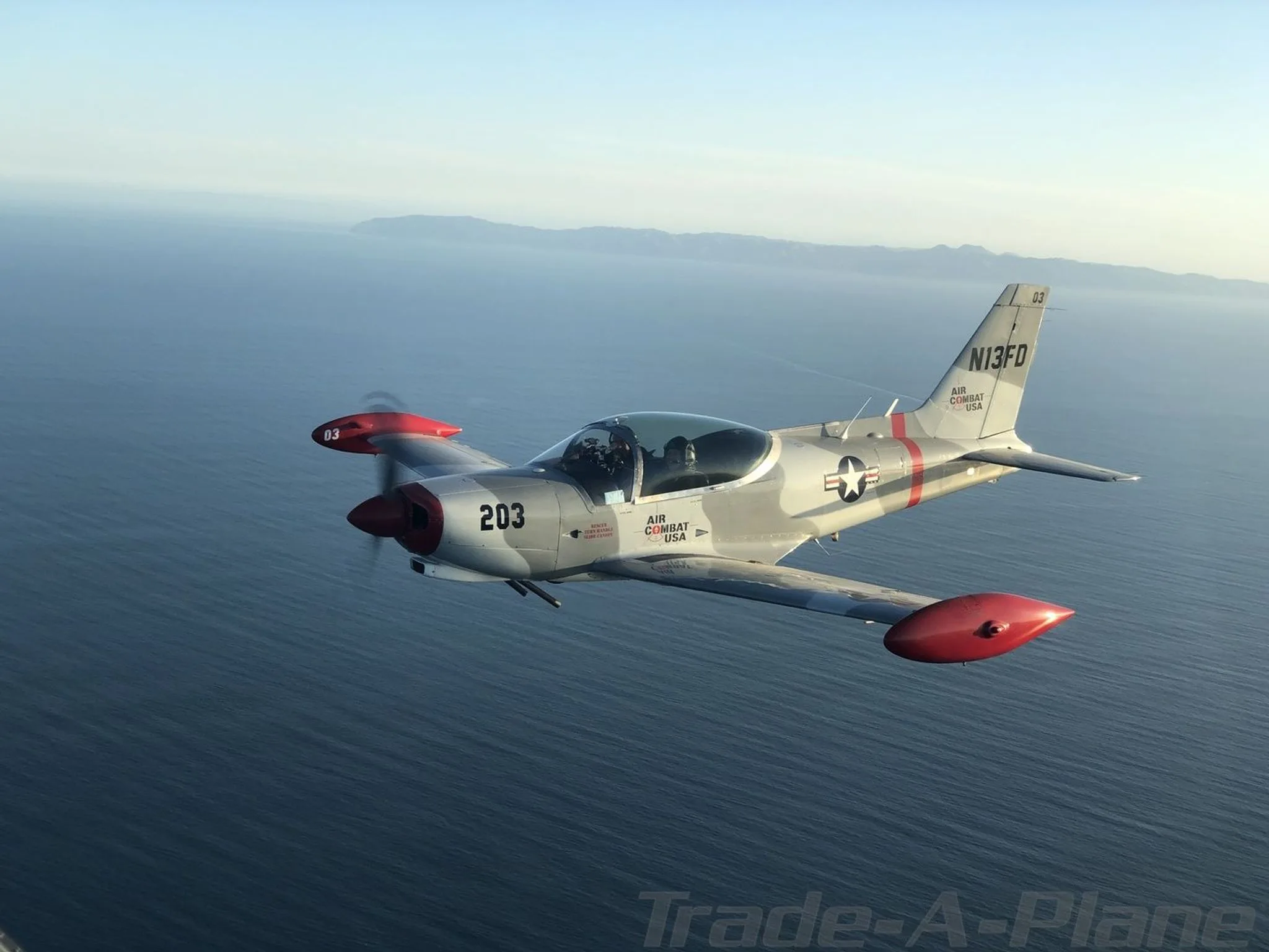 A gray and red single-engine aircraft, N13FD, with 'Air Combat USA' markings, flies over the ocean.
