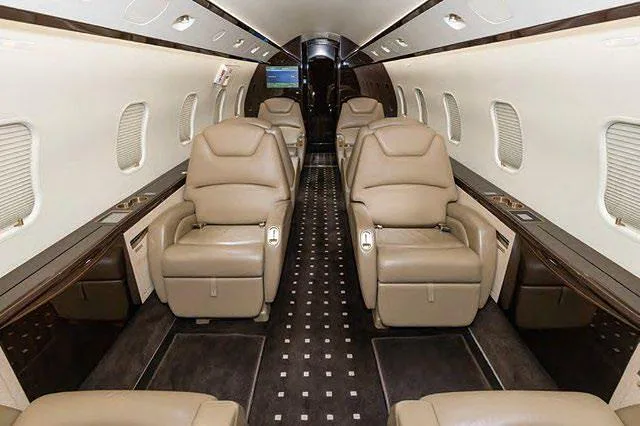 The luxurious interior cabin of a business jet with two rows of opposing beige leather seats and dark patterned carpet.