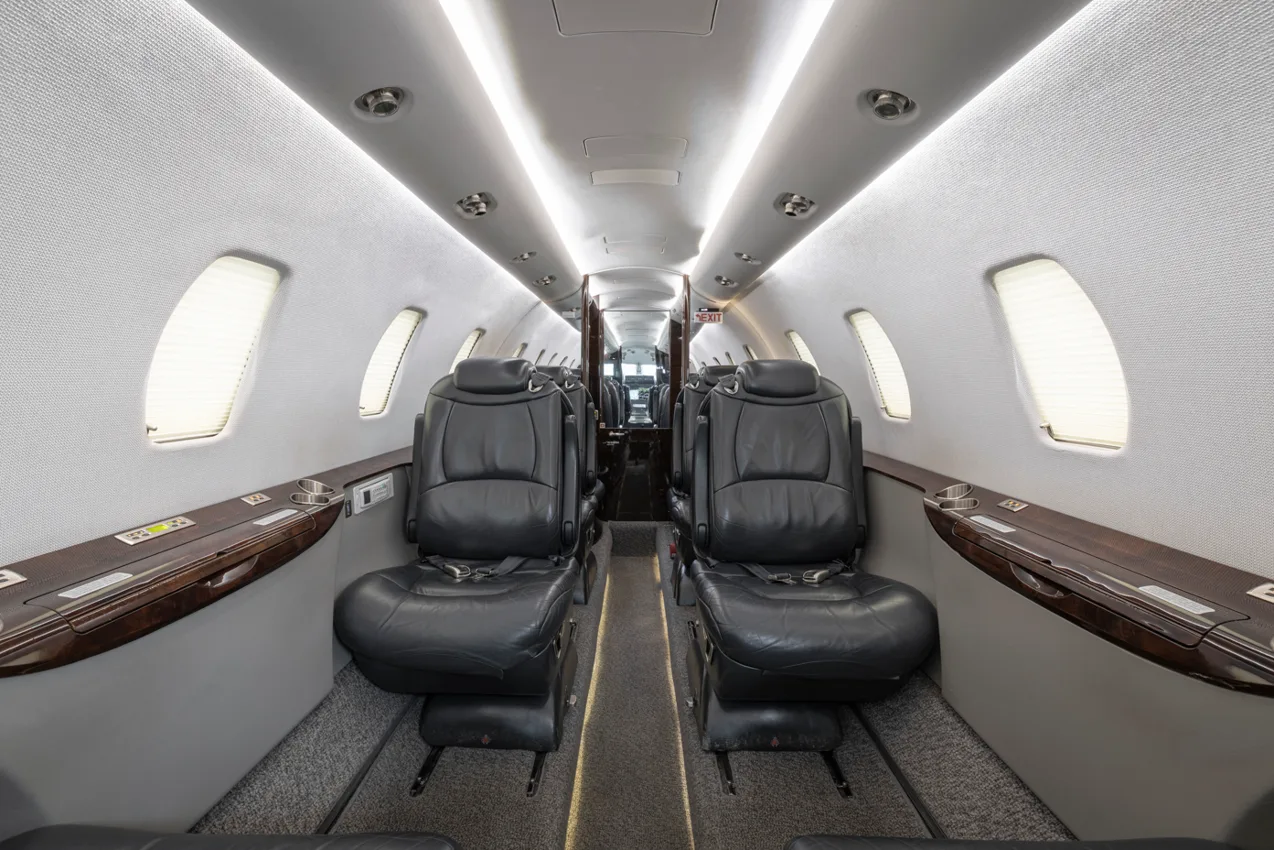 The interior cabin of a business jet, showing rows of black leather seats and overhead lighting.