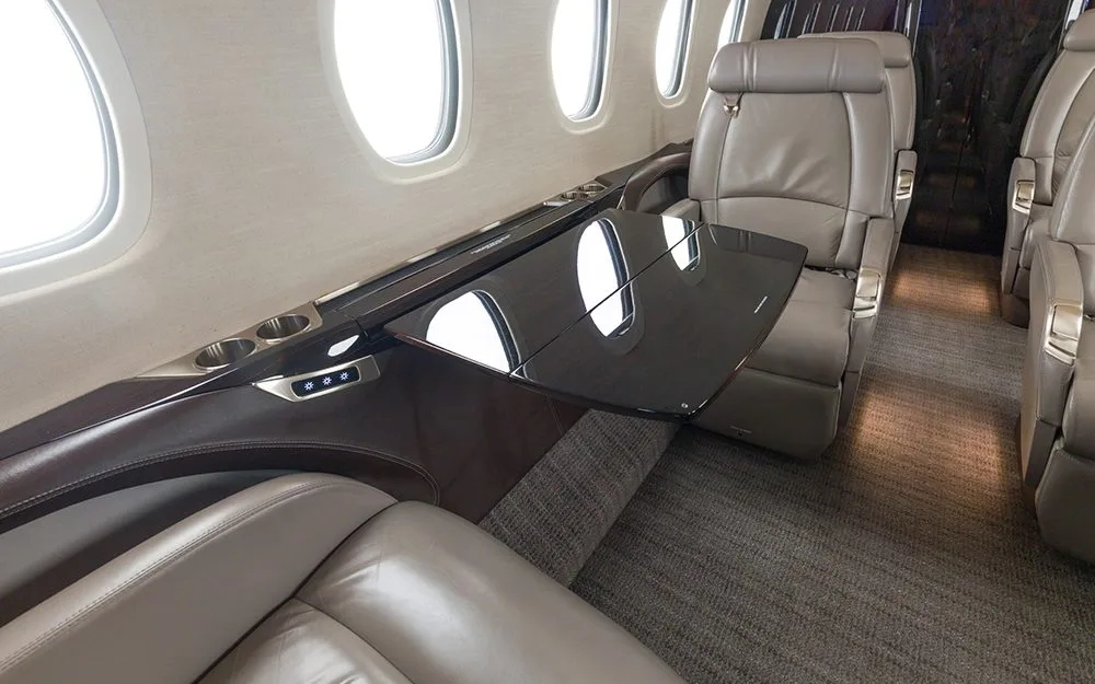 A close-up view of a light brown leather seat and a deployed black fold-out table in a private jet cabin.